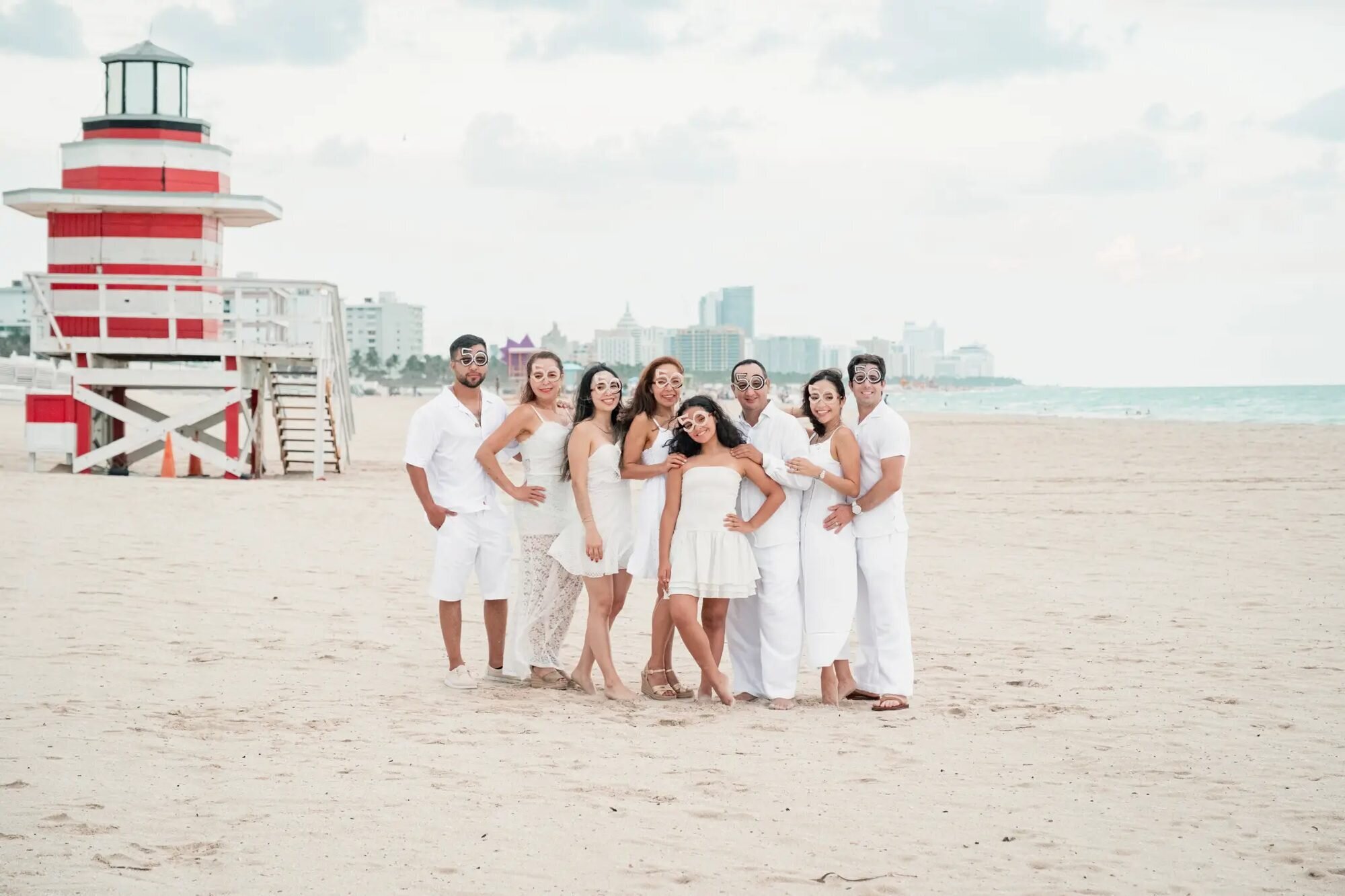 A large family of eight dressed in white laughing together on the sand at South Beach, Miami, with the iconic red-and-white lifeguard tower in the background — captured by a Dreamgraphy Miami family photographer