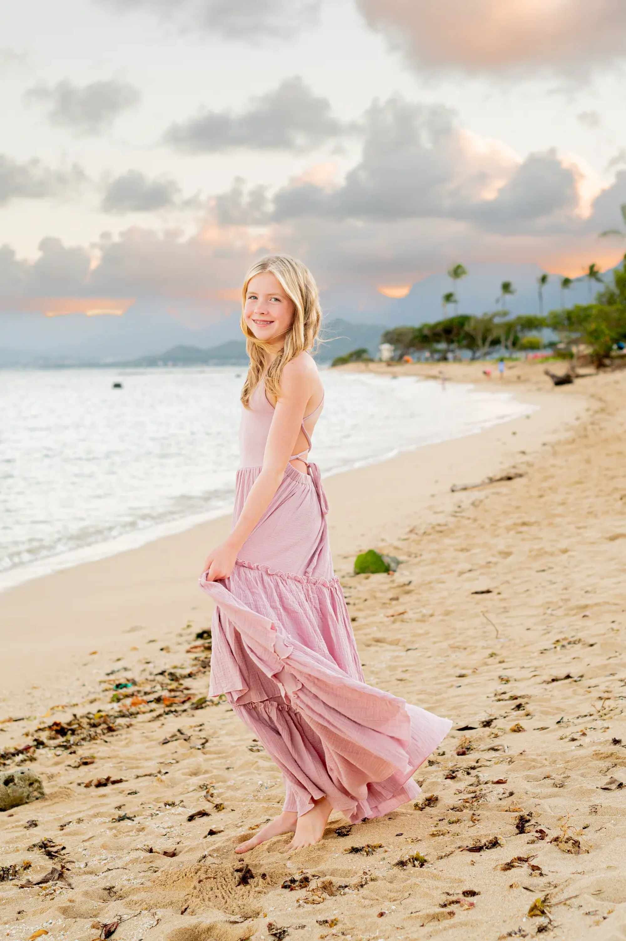 A young girl in a flowing pink dress twirling on a sandy Kauai beach at golden hour, with soft waves and a pastel sunset sky in the background, captured during a professional family photo session