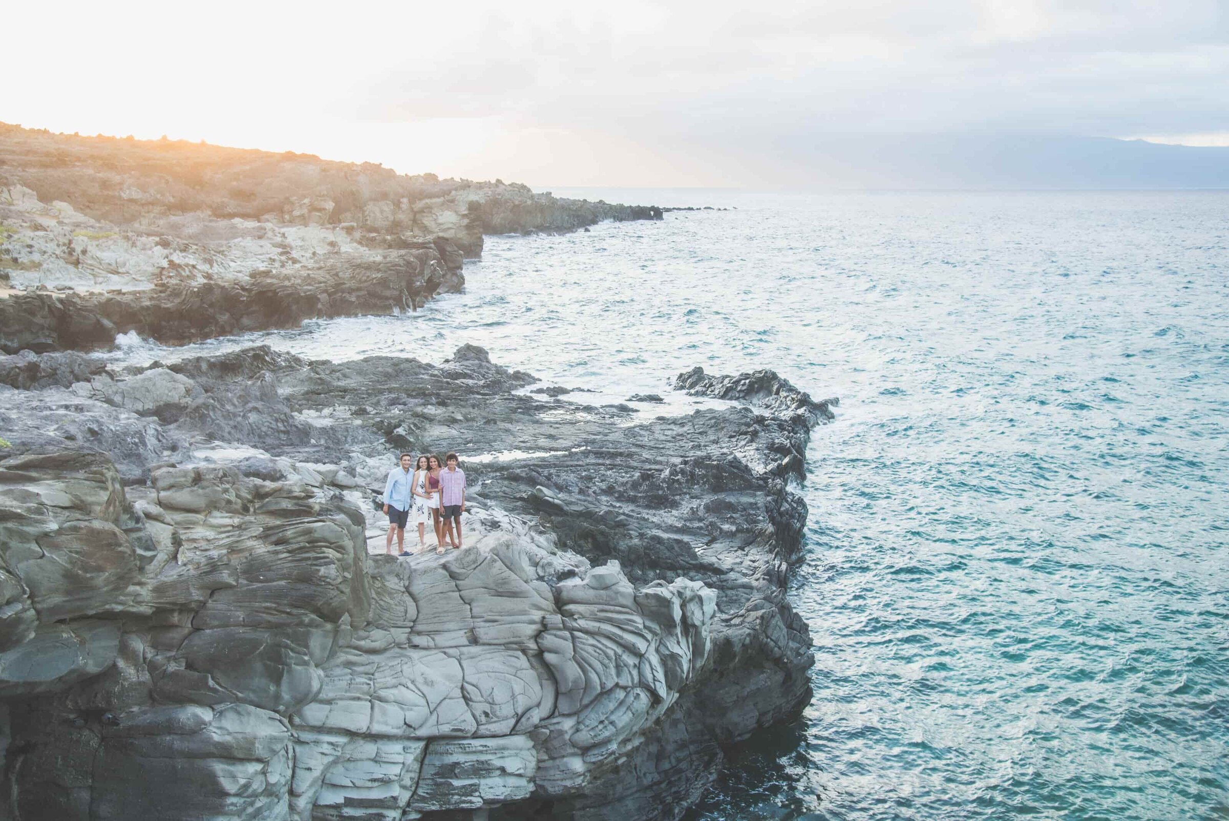 ironwood-beach-maui A family of four standing together on wide grey lava-rock cliffs above the ocean, with the sun low over the headland, at Ironwood Beach on Maui