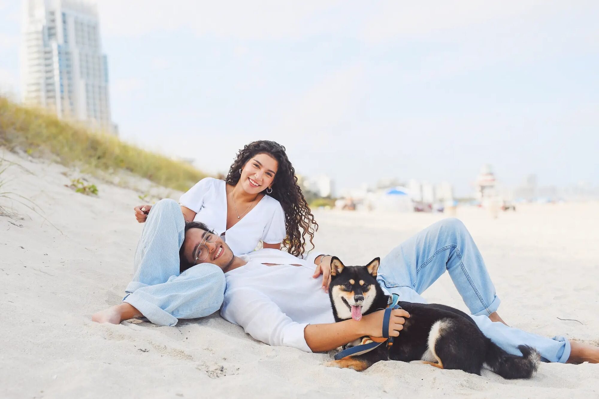 A couple relaxing on the sand dunes at South Beach, Miami, with their Shiba Inu dog, with the Miami skyline in the background — captured by a Dreamgraphy Miami couple photographer