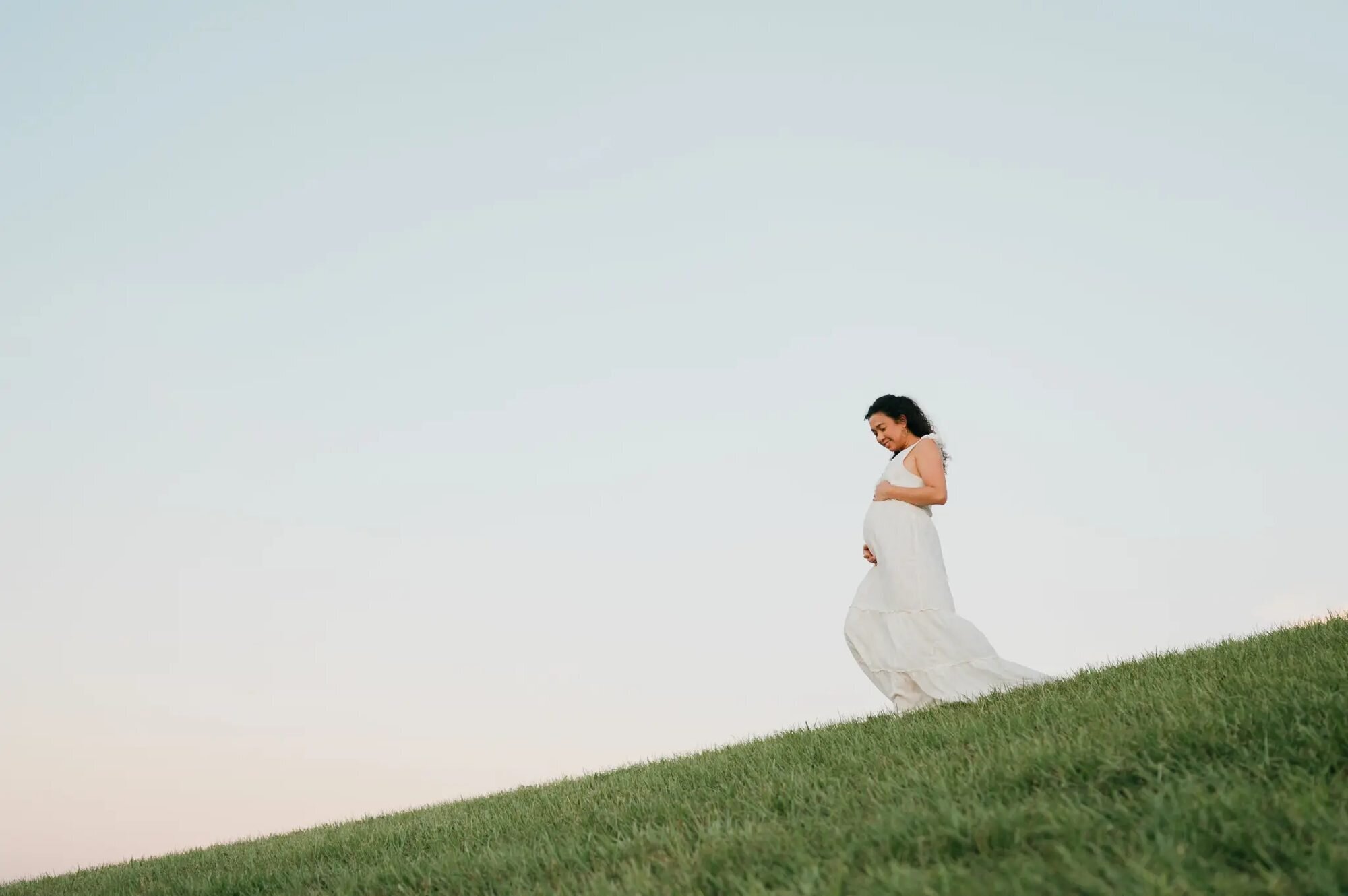 A pregnant woman in a flowing white dress cradling her baby bump while standing on a grassy hillside at golden hour in Tampa, Florida — captured by a Dreamgraphy Tampa maternity photographer