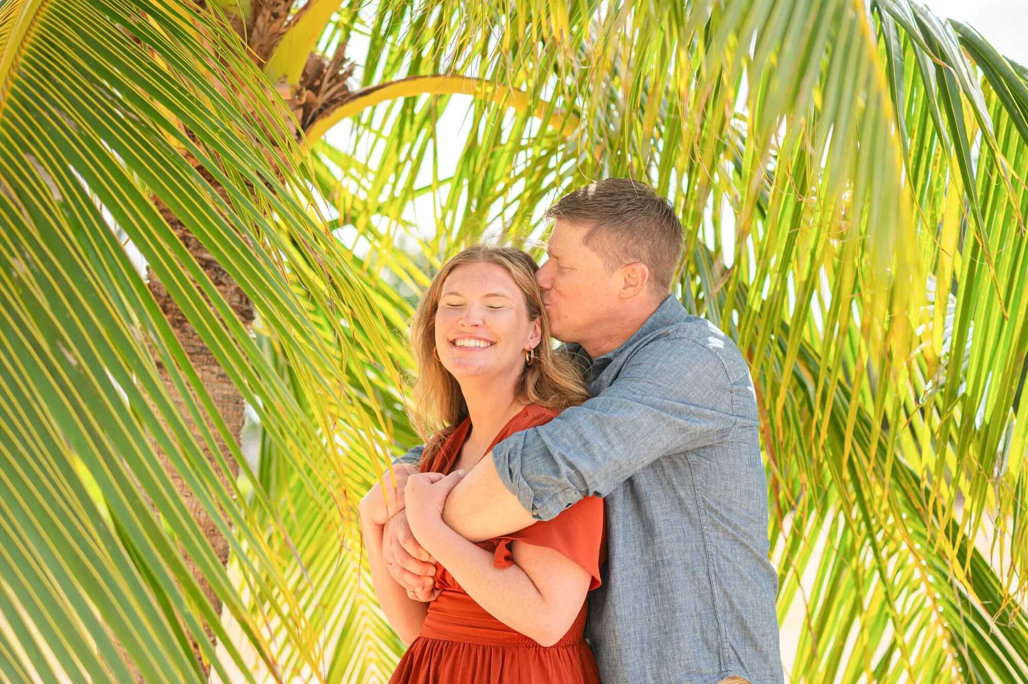 A man kissing a laughing woman on the cheek as they embrace beneath lush tropical palm fronds during a couples photography session on Oahu