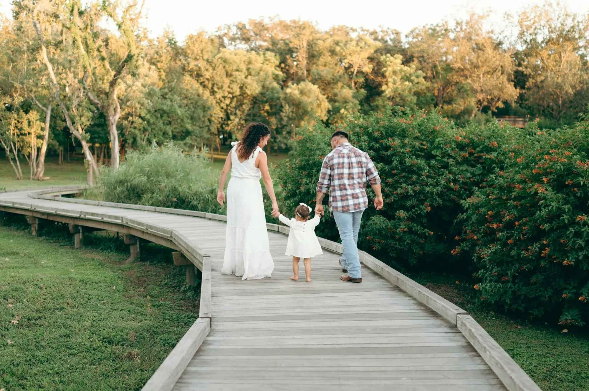 Orlando family photographer capturing a family of three walking hand-in-hand along a winding wooden boardwalk through a lush Florida park at golden hour