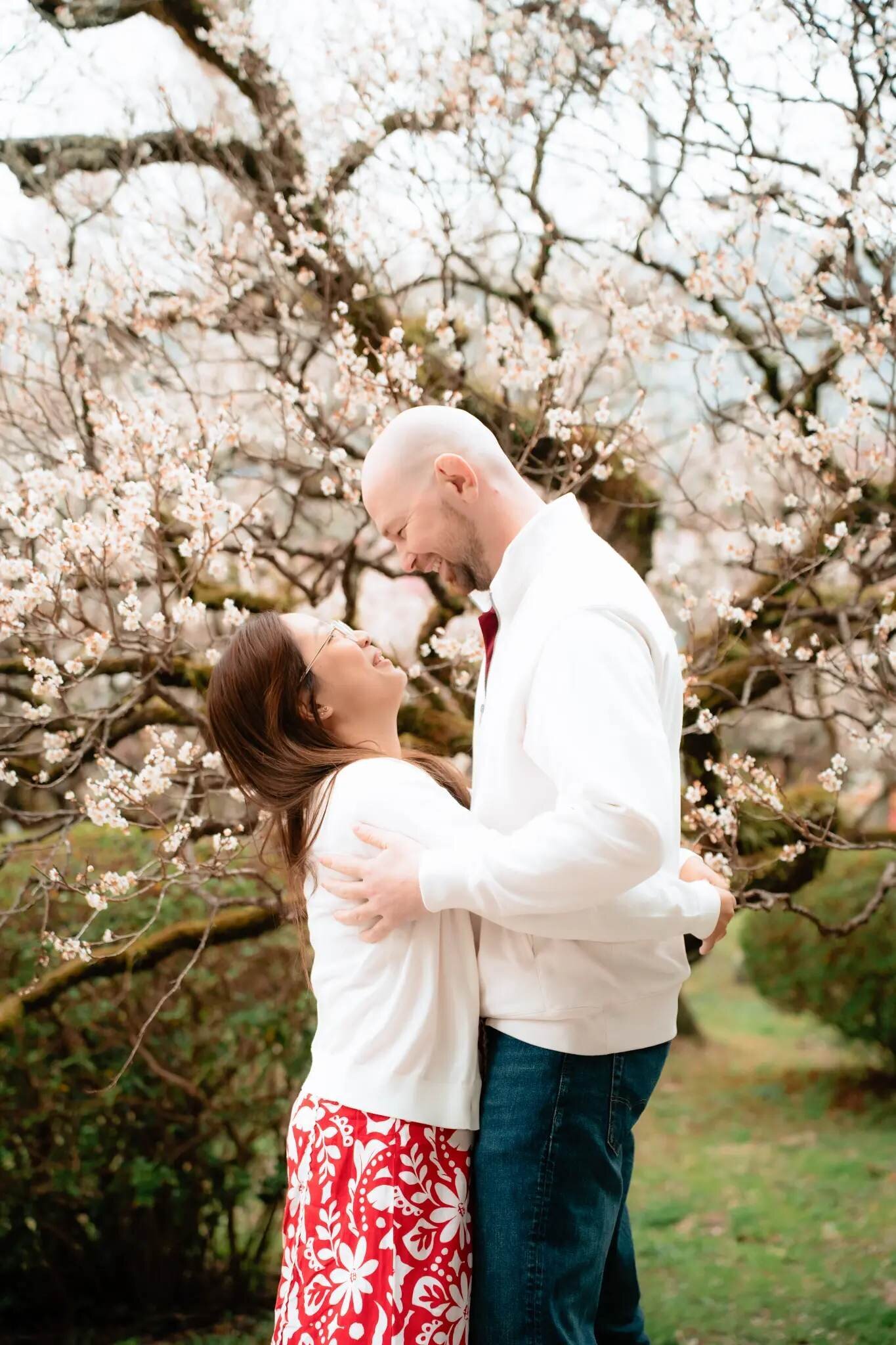A laughing couple in white tops embraces beneath a tree covered in pale blossoms at Uminonakamichi Seaside Park in Fukuoka.