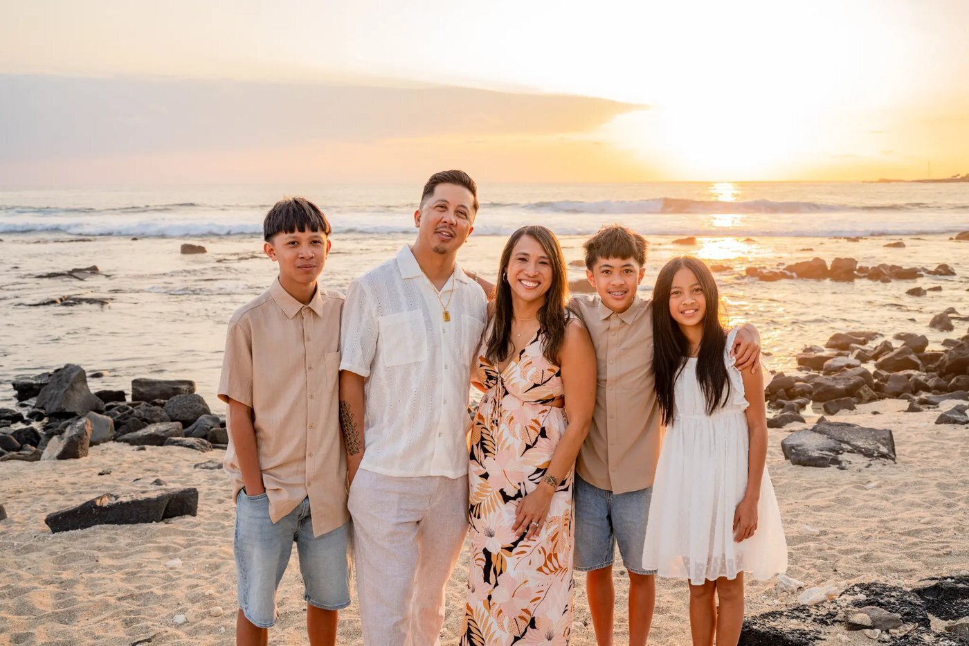 A family of five smiling together on a sandy Big Island beach at golden sunset with lava rocks and ocean waves behind them