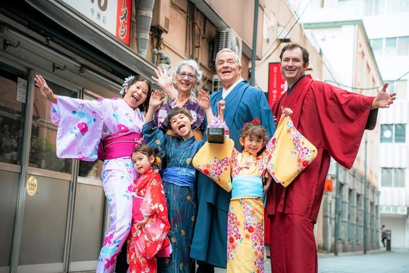 Seven adults and children in colorful kimono cheer and wave on a narrow street lined with shop signs in Nakasu, Fukuoka.
