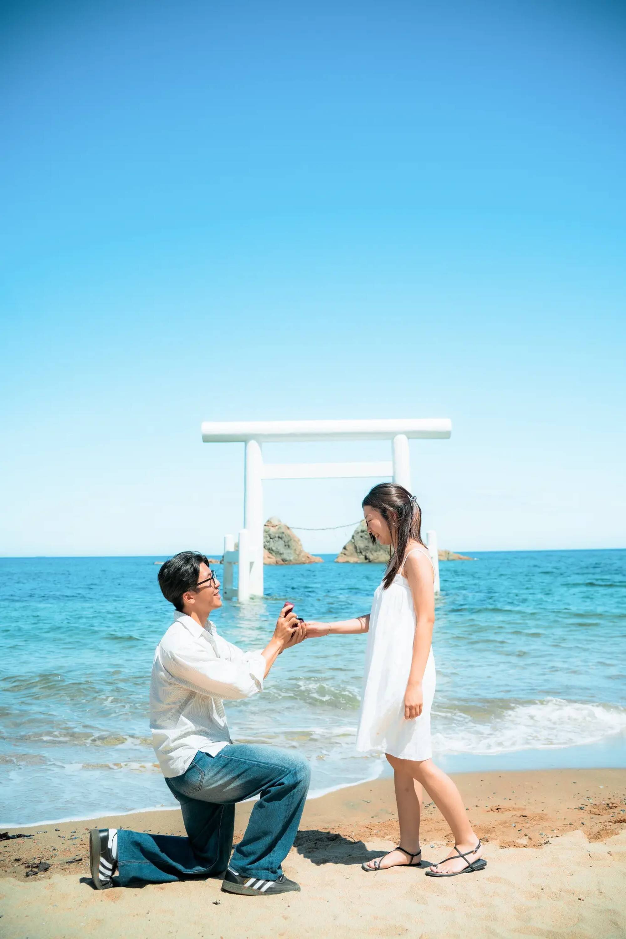 A man on one knee offers a ring to a smiling woman in a white sundress on a beach in Fukuoka, with a white torii gate standing in the sea
