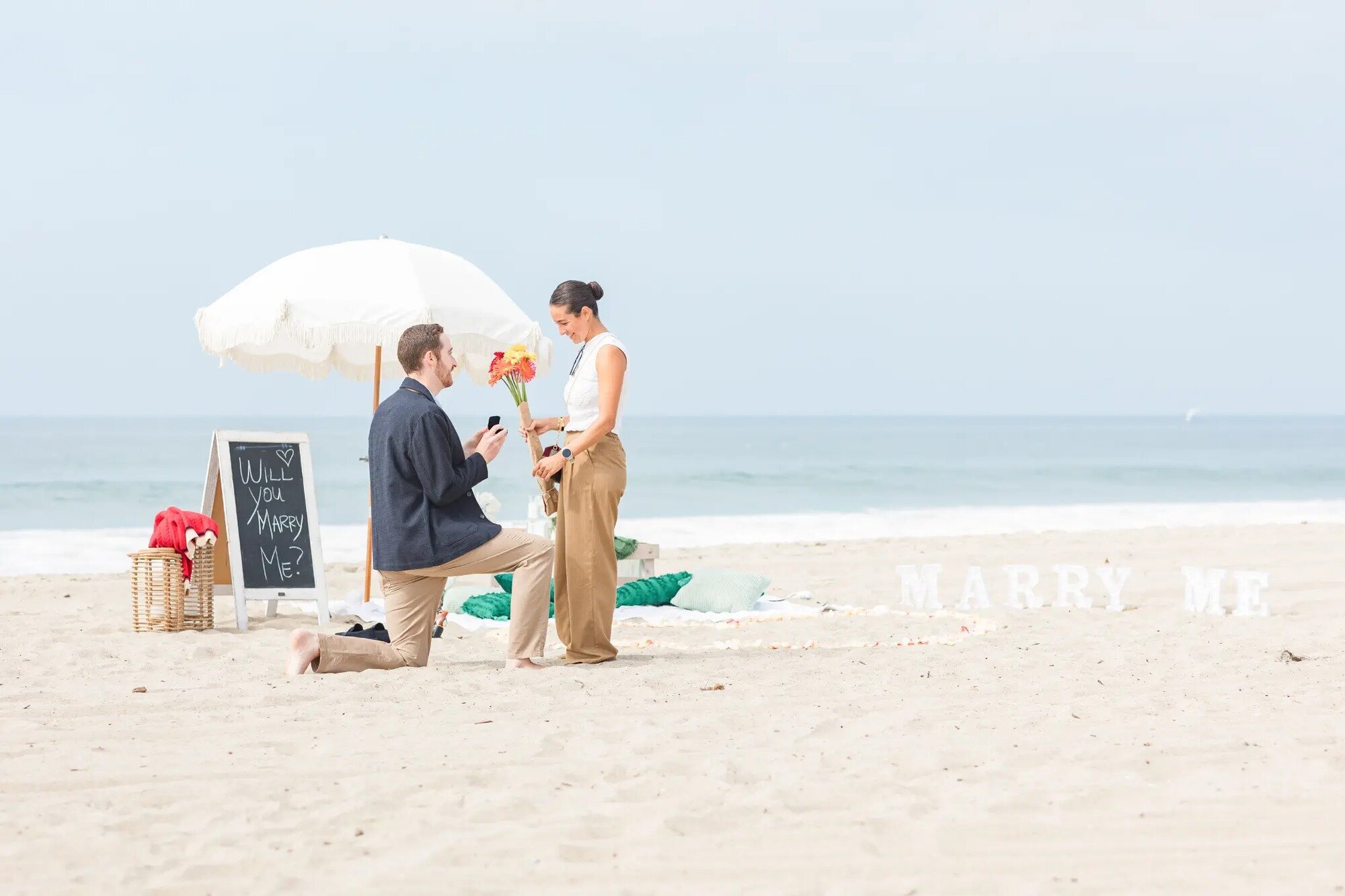Los Angeles Photographer — Surprise Proposal Session at Santa Monica Beach Los Angeles photographer capturing a man proposing on one knee to his partner on Santa Monica Beach with a "Will You Marry Me" sign and floral setup