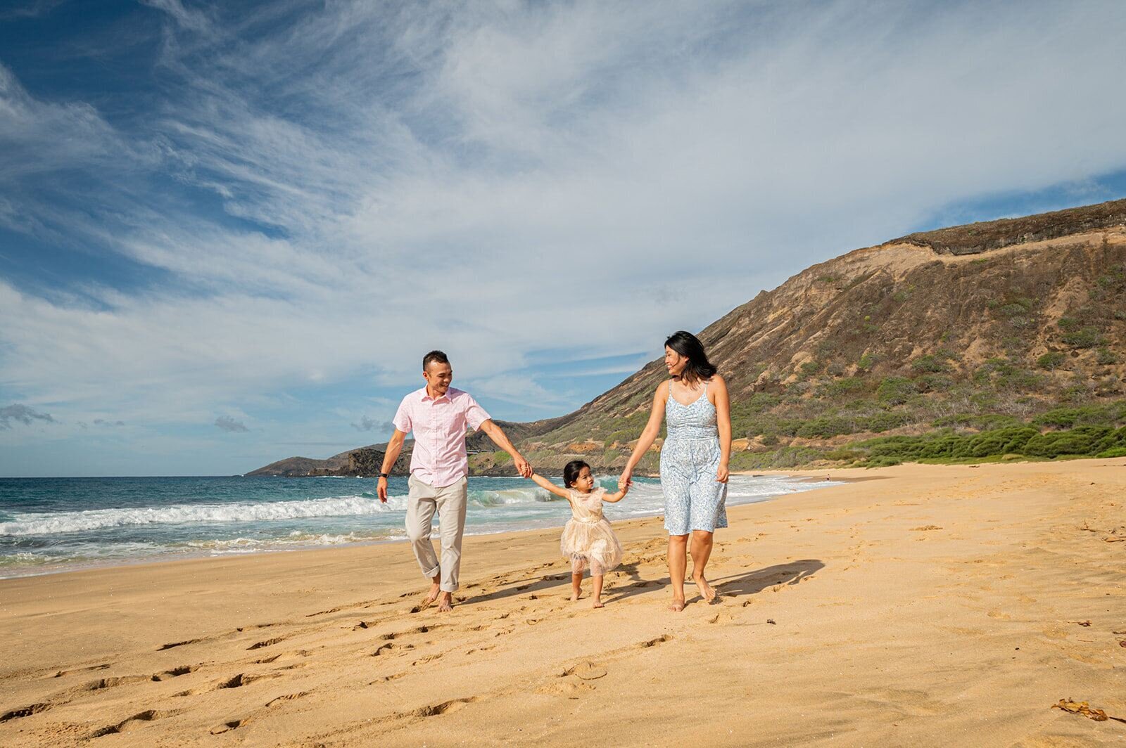A family of three holding hands and walking along Sandy Beach Park on Oahu with dramatic golden cliffs and crashing waves in the background