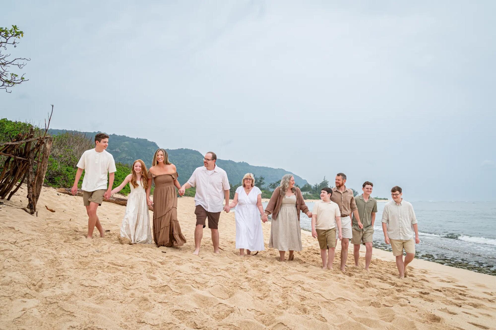 A large multigenerational family holding hands and walking together along a sandy Kauai beach, with lush green cliffs and the ocean in the background, captured during a professional family photo session