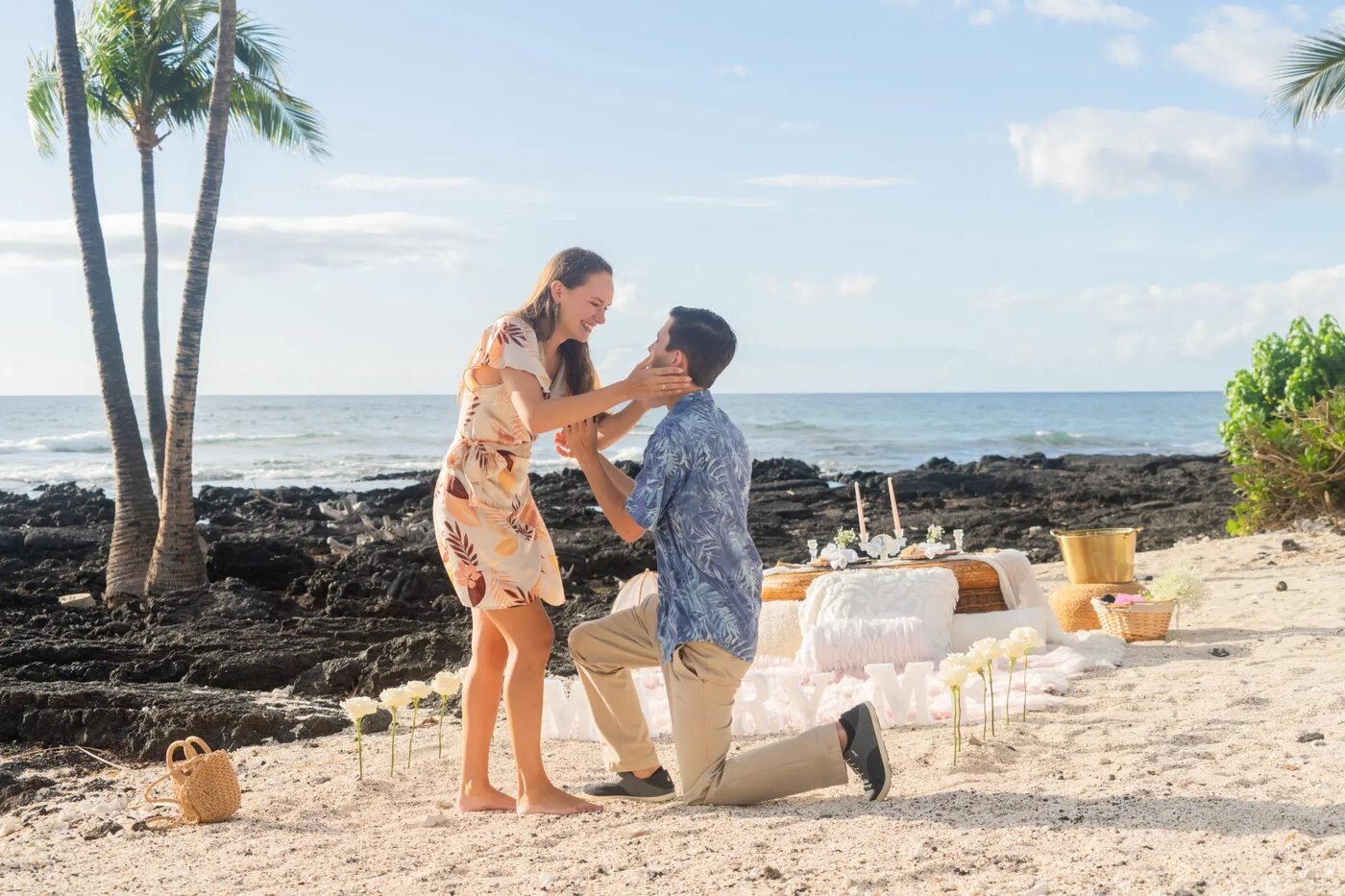 A man proposing on one knee to his partner on a sandy Big Island beach with lava rocks and palm trees in the background