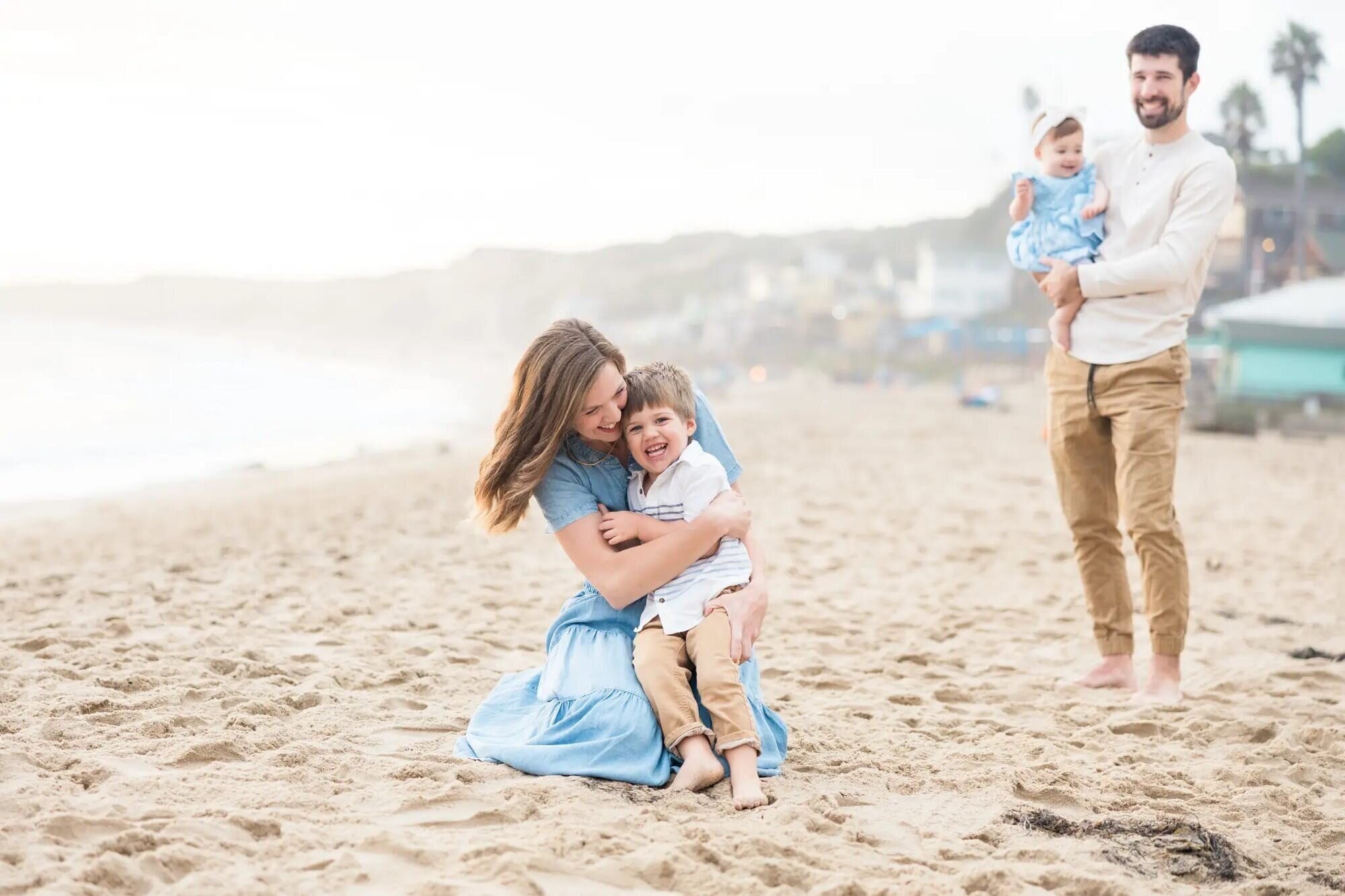 Mother hugging her young son on the sand while father holds baby during a candid Los Angeles family photography session at Santa Monica Beach