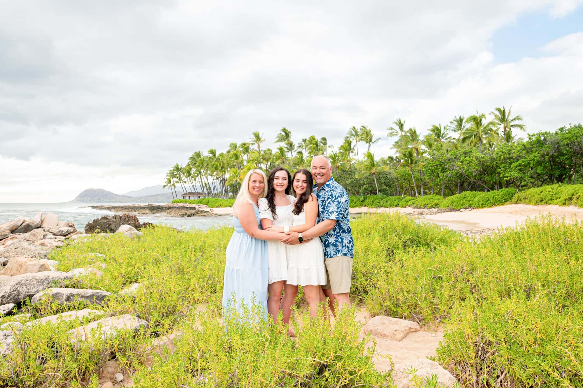 A family of four smiling and embracing on a rocky Hawaii coastline surrounded by lush greenery and palm trees during a professional vacation photography session