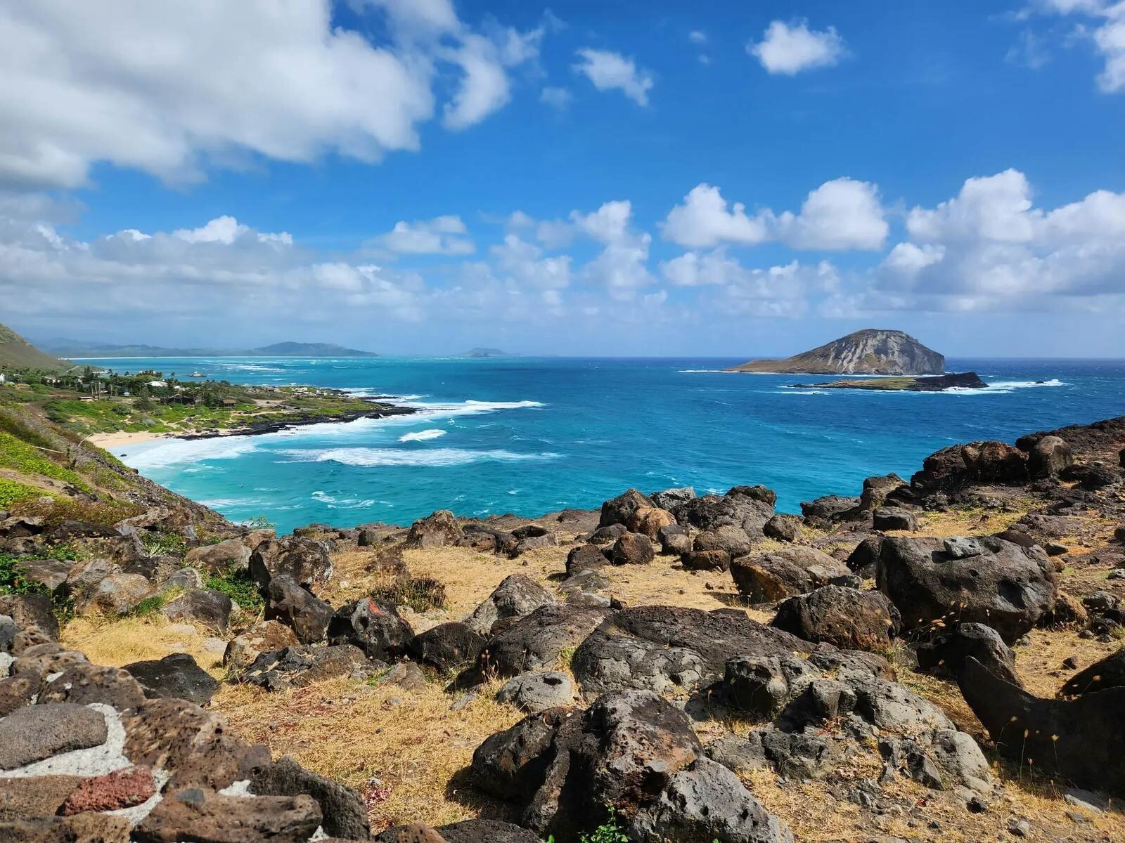Dark volcanic boulders and dry grass above a turquoise bay with white surf, a sandy beach and a steep offshore island in Oahu