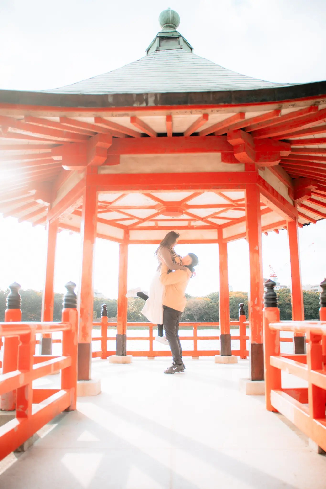A couple sharing a joyful lift inside the iconic red pavilion at Ohori Park in Fukuoka, surrounded by soft natural light and the park's serene lakeside setting. Photographed by Dreamgraphy.
