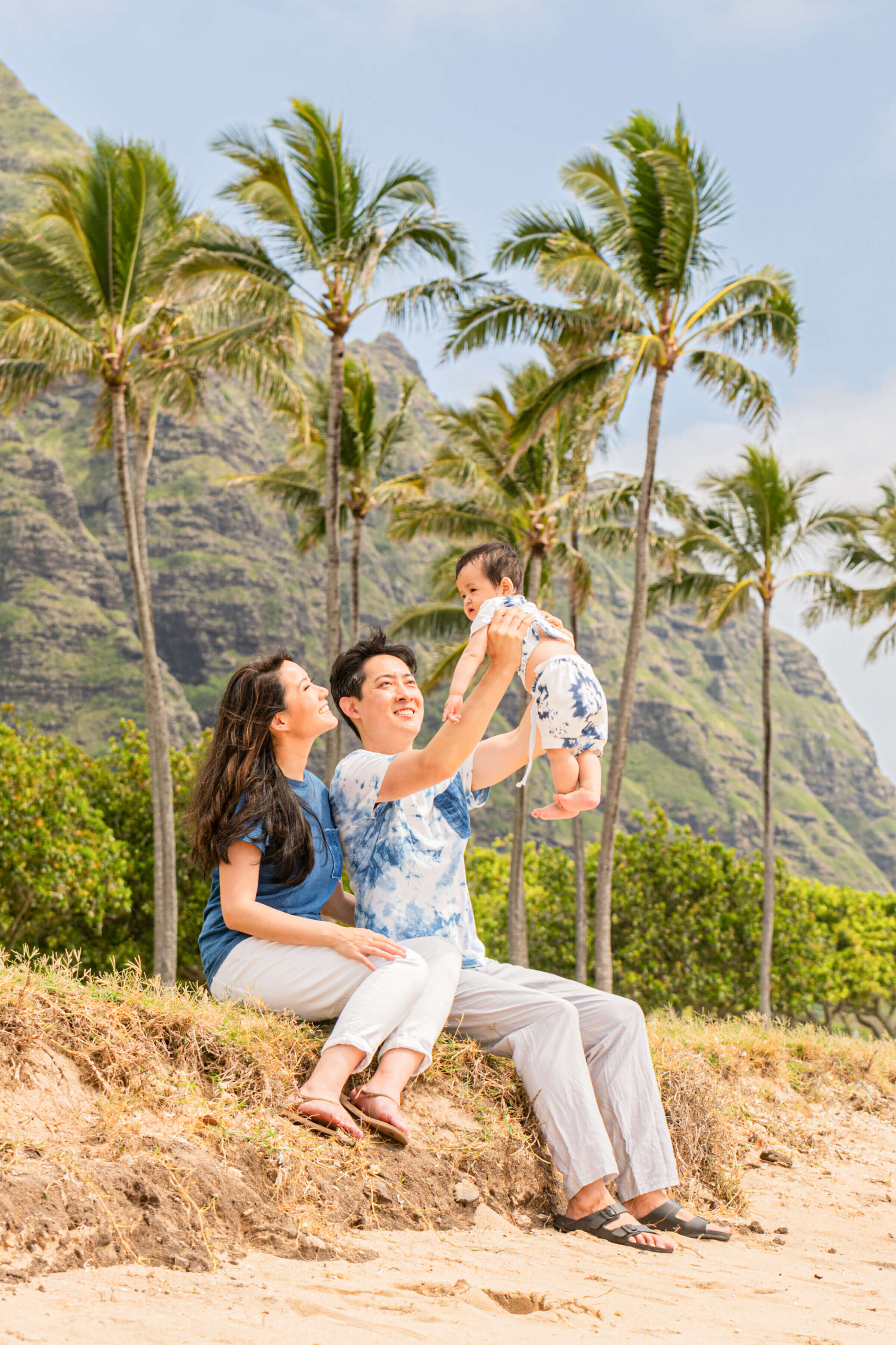 A couple sitting on a sandy hillside lifting their baby in the air and laughing during a family vacation photography session in Hawaii, with tall palm trees and dramatic green mountain cliffs in the background