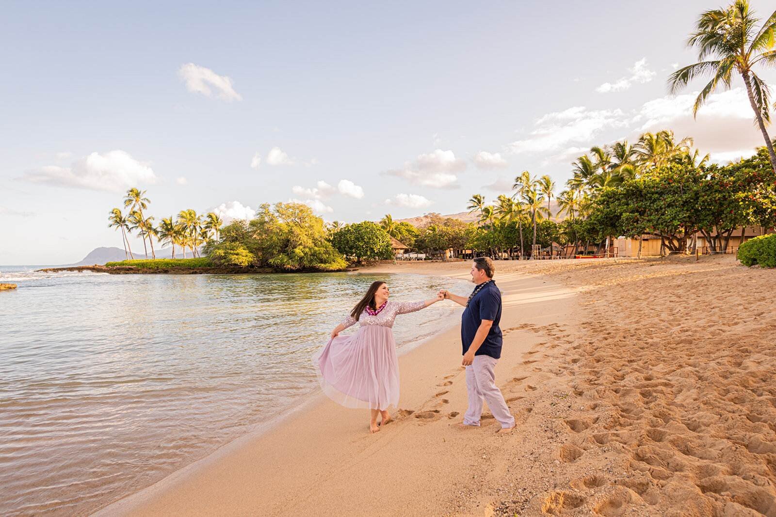 A couple dancing and spinning on the sandy shore at Ko Olina Beach on Oahu at golden hour with calm water, and lush palm trees,