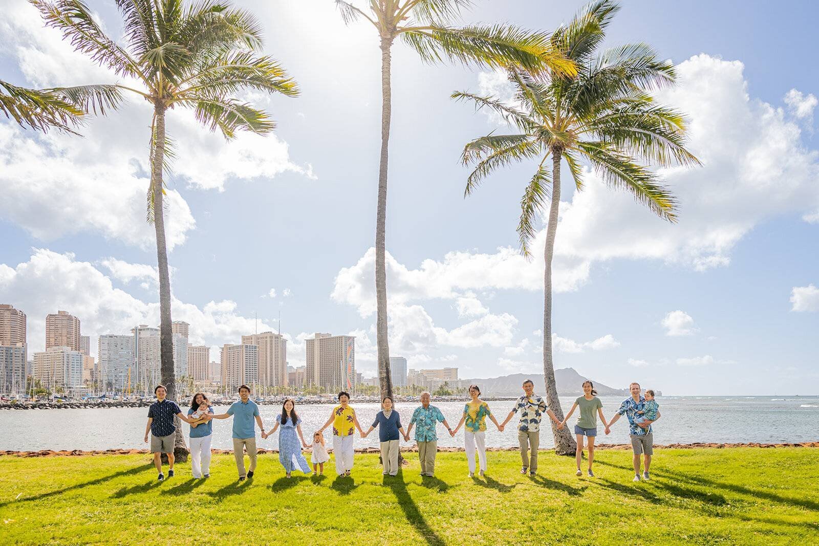 Oahu Photographer — Extended Family Session at Ala Moana Beach Park A large extended family of twelve holding hands in a line on the grass at Ala Moana Beach Park with tall palm trees, the Waikiki skyline, and Diamond Head in the background