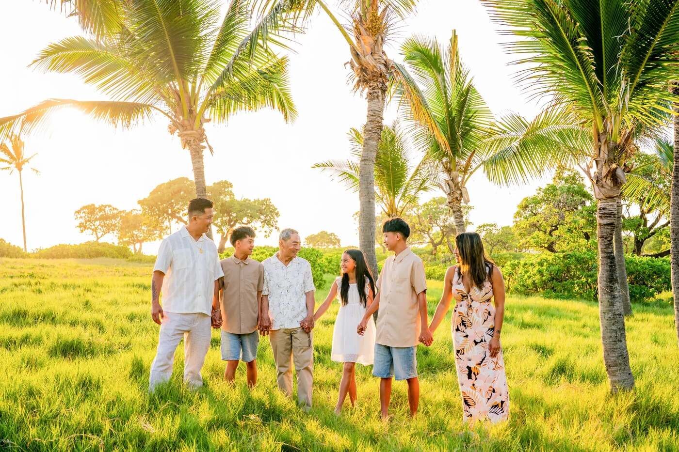 An extended family of six holding hands and walking together through lush green grass under tall palm trees on the Big Island of Hawaii during golden hour