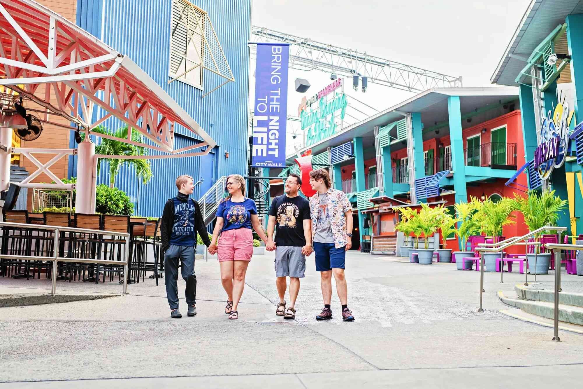 A family of four holding hands and walking together through the colorful, vibrant promenade of Universal CityWalk in Orlando, Florida, with bright tropical-themed buildings and a CityWalk banner in the background
