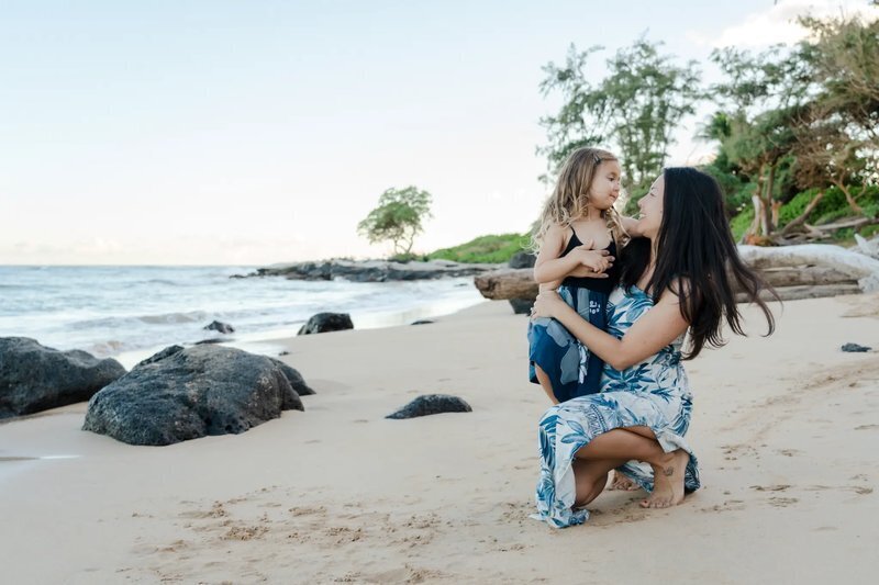 A mother crouching on a sandy Kauai beach holding her young daughter and smiling during a professional family photo session, with ocean waves and tropical trees in the background