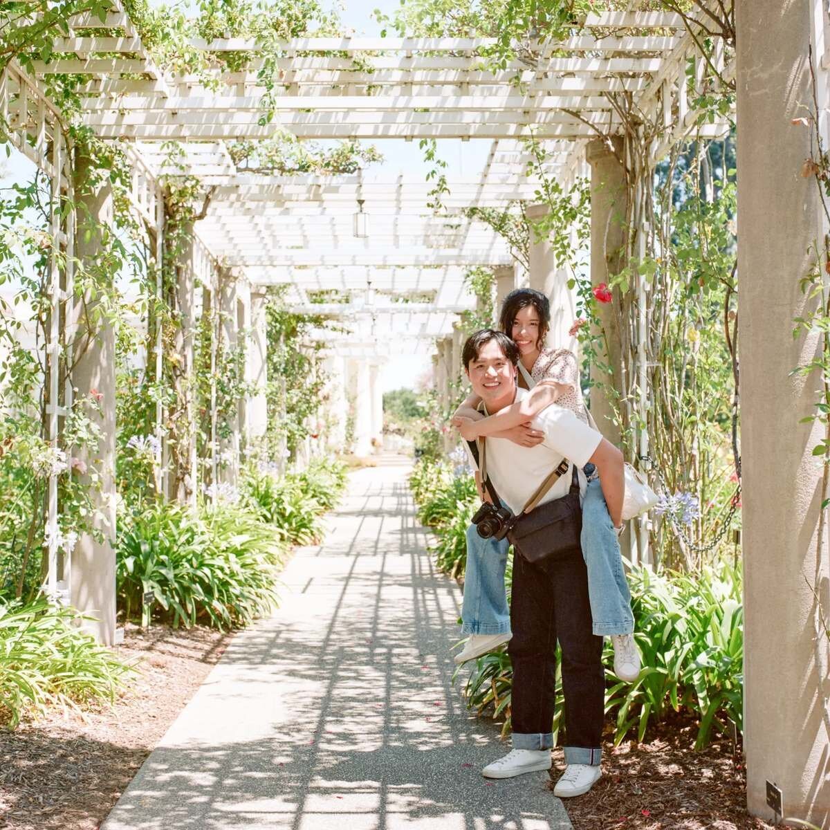 Los Angeles couples photographer capturing a couple laughing together as the man gives his partner a piggyback ride under a white pergola covered in climbing roses at the Huntington Library Rose Garden