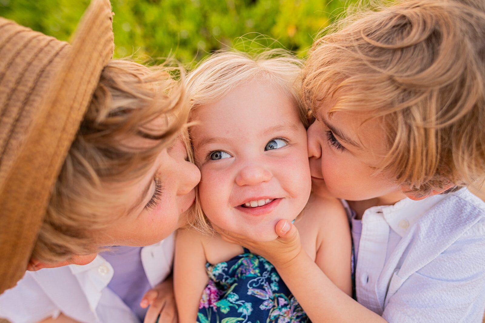 Two older siblings kissing a laughing toddler on both cheeks simultaneously during a family photography session in Hawaii with lush green foliage in the background
