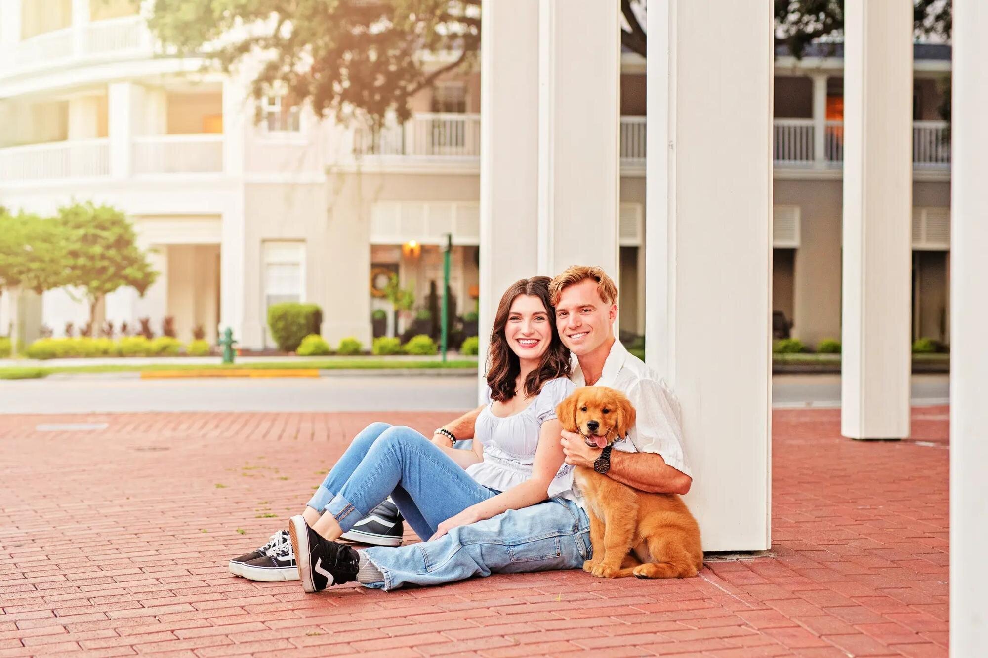 A smiling couple sitting on a brick sidewalk in downtown Tampa with their golden retriever puppy, surrounded by white colonial-style architecture — captured by a Dreamgraphy Tampa couple photographer