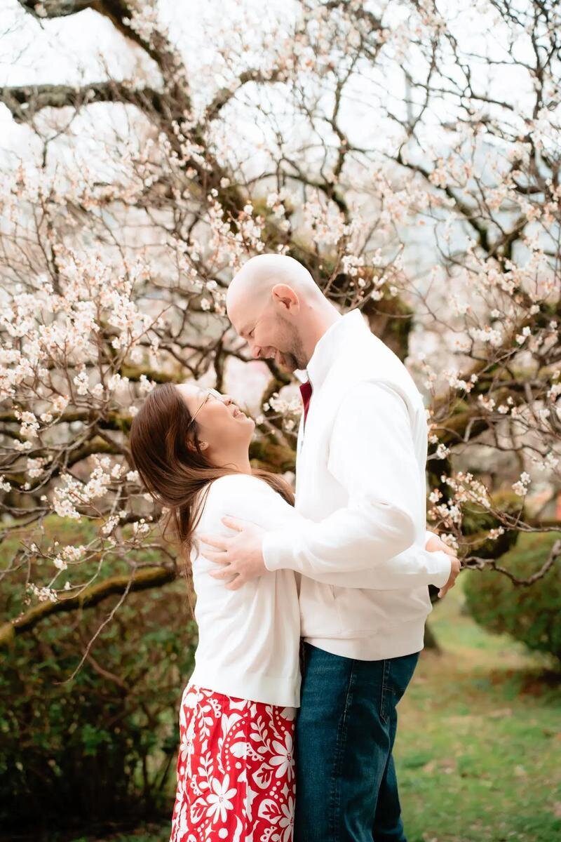 A laughing couple in white tops embraces beneath a tree covered in pale blossoms at Uminonakamichi Seaside Park in Fukuoka.