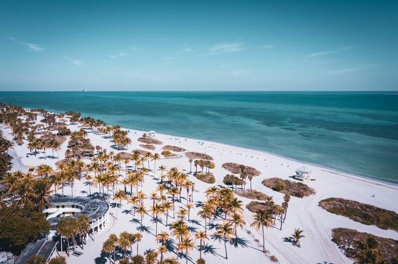 Aerial view of a white-sand beach dotted with palm trees, a lifeguard stand and a curved pavilion beside turquoise water in Miami