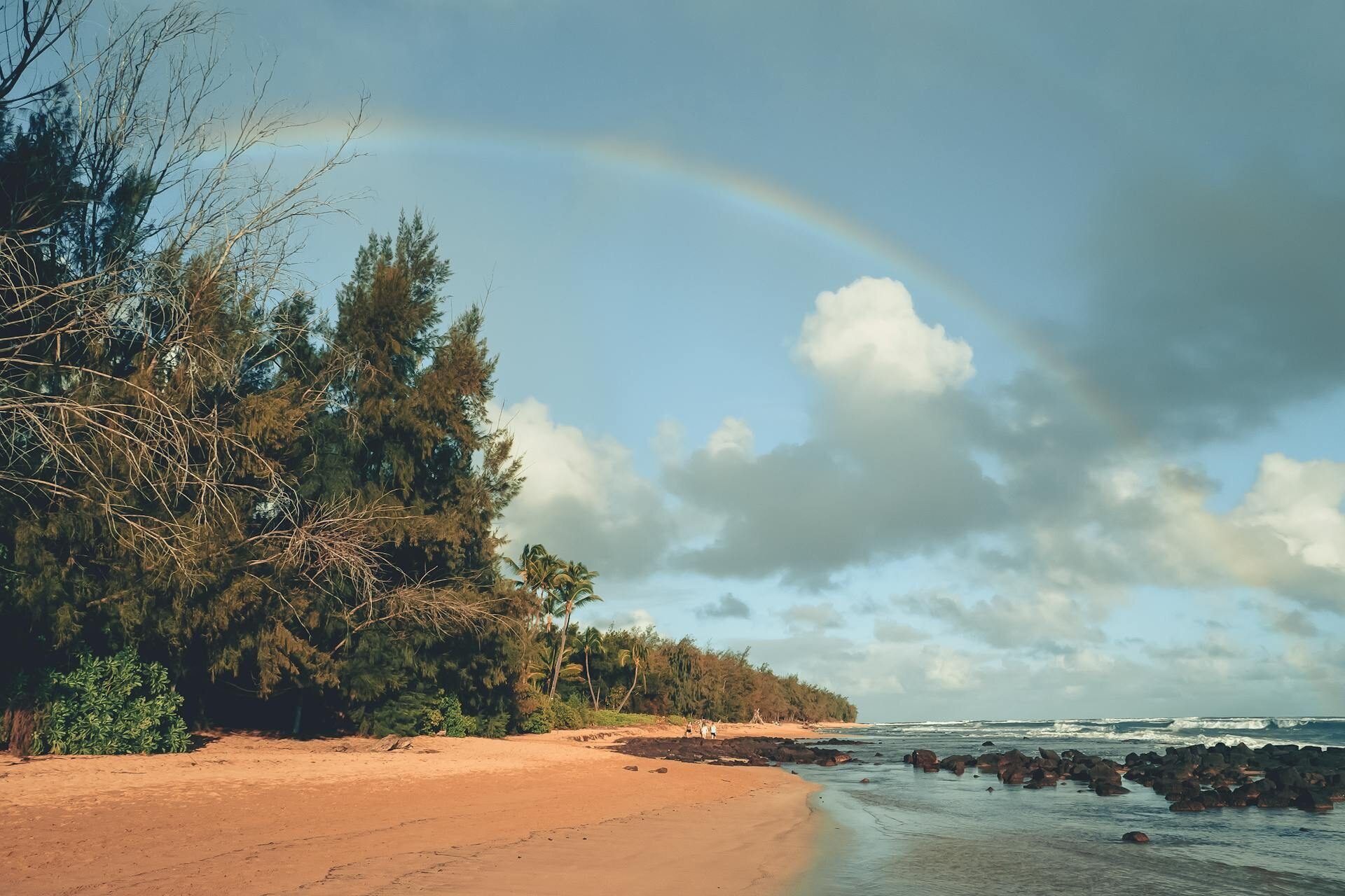 A rainbow arches over a sandy beach lined with trees and palms, with dark rocks in the shallows and three distant walkers in Kauai