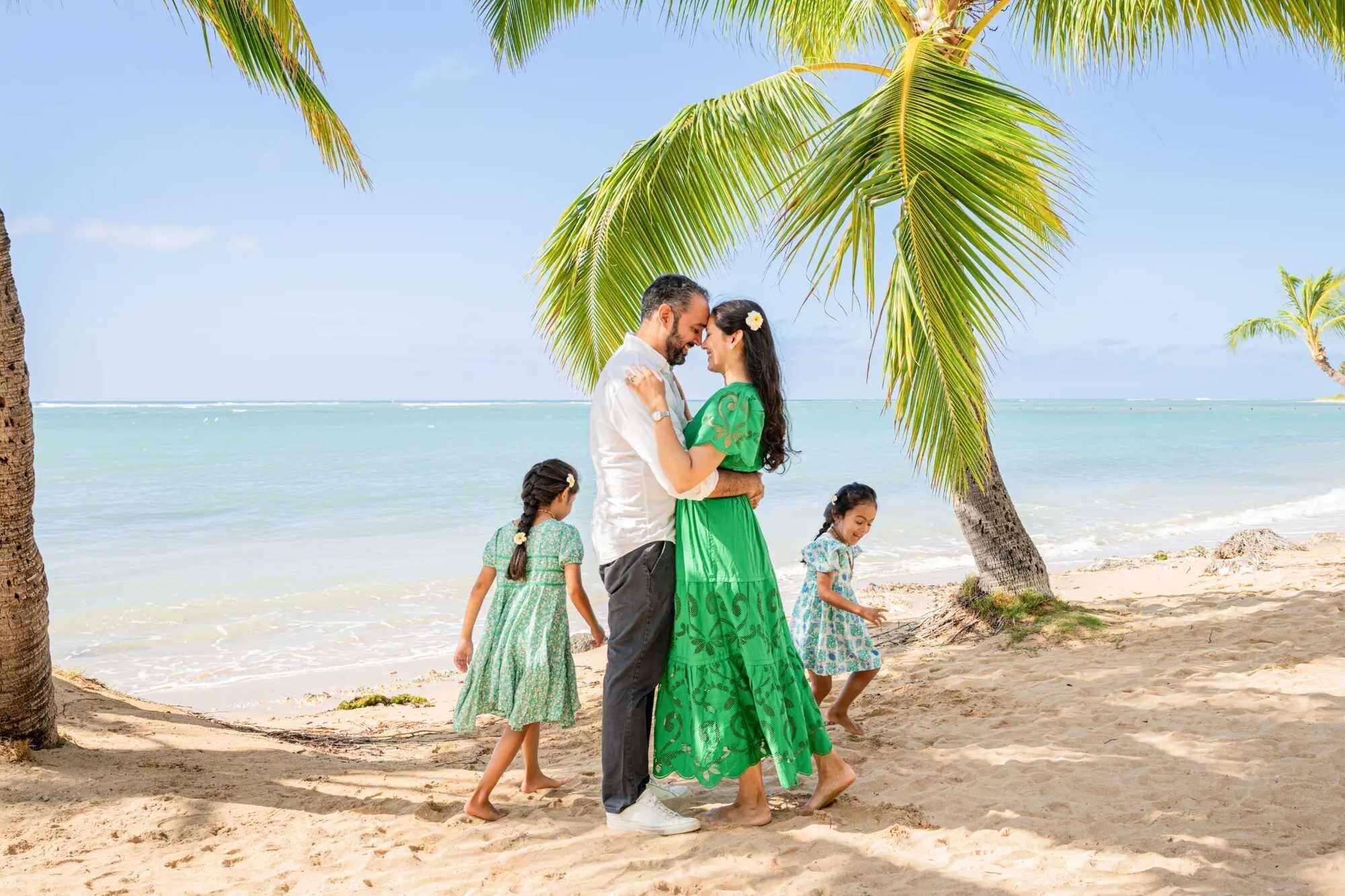 A couple touching foreheads and embracing under a leaning palm tree on Kahala Beach on Oahu while their two young daughters play in the sand behind them with turquoise water in the background