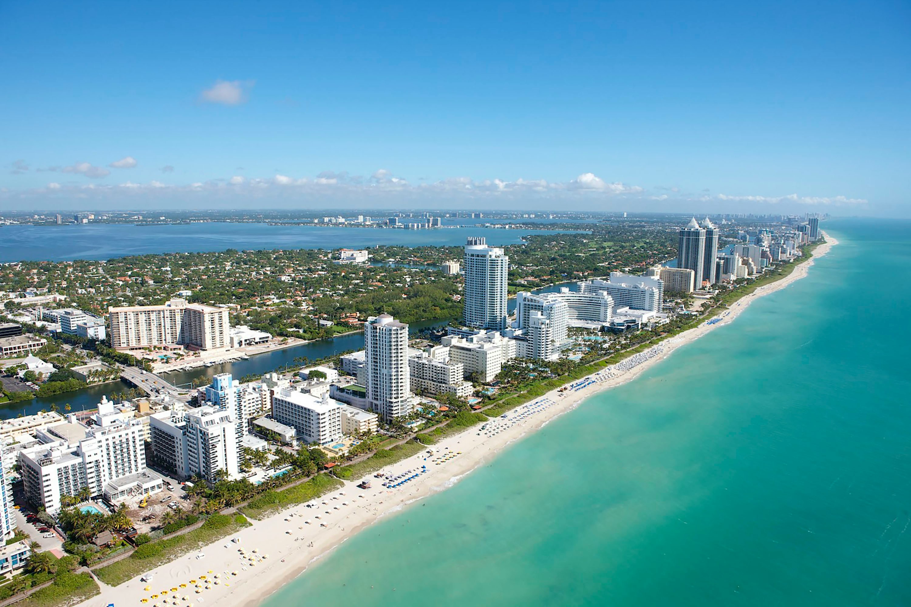 Miami Beach aerial view: the beach, the hotels and Biscayne Bay Aerial view of Miami Beach, with white high-rise hotels along a wide sandy beach, turquoise Atlantic water, and Biscayne Bay in the distance