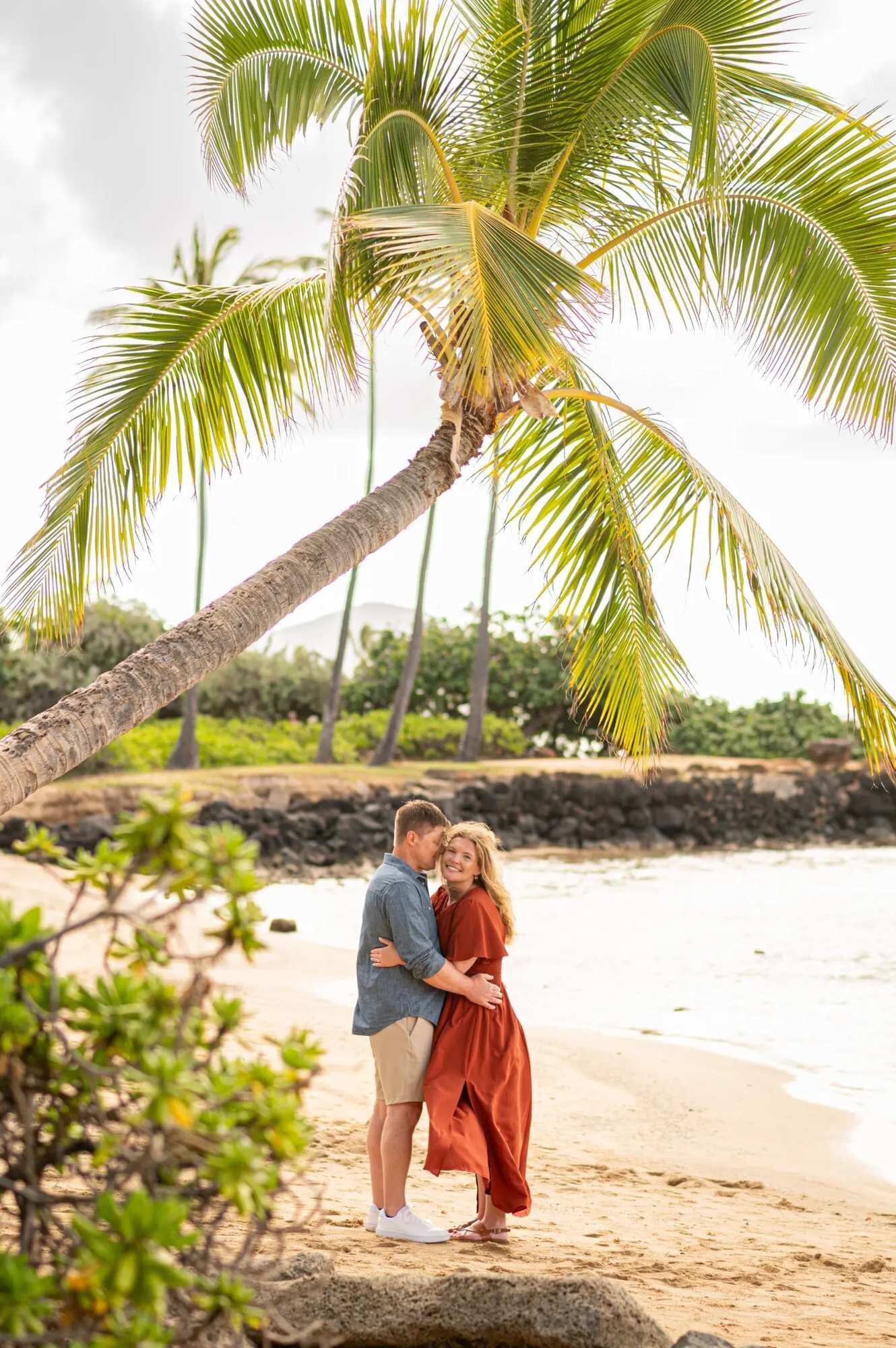 Couples Photographer Oahu Hawaii — Romantic Beach Portrait Under Palm Tree A couple embracing on a sandy Oahu beach under a tall leaning palm tree with calm ocean water and lush greenery in the background