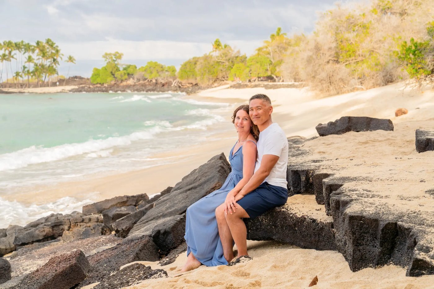 A couple sitting close together on lava rocks on the Big Island of Hawaii with turquoise ocean waves and a white sand beach curving behind them