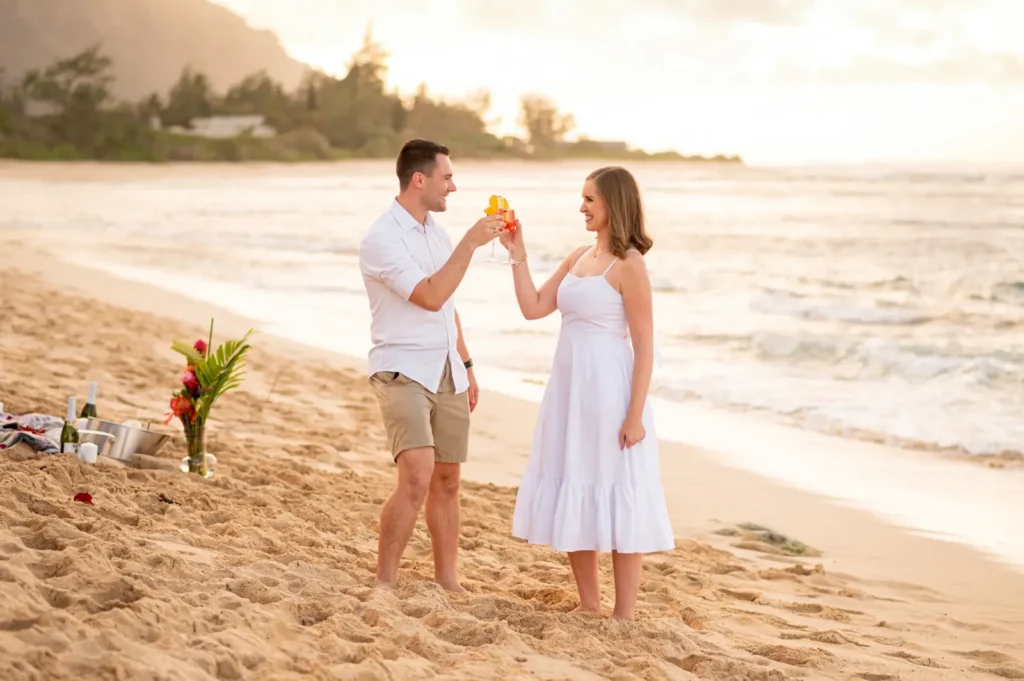 Couple celebrating a beach proposal in Hawaii with champagne and a flower arrangement at sunset