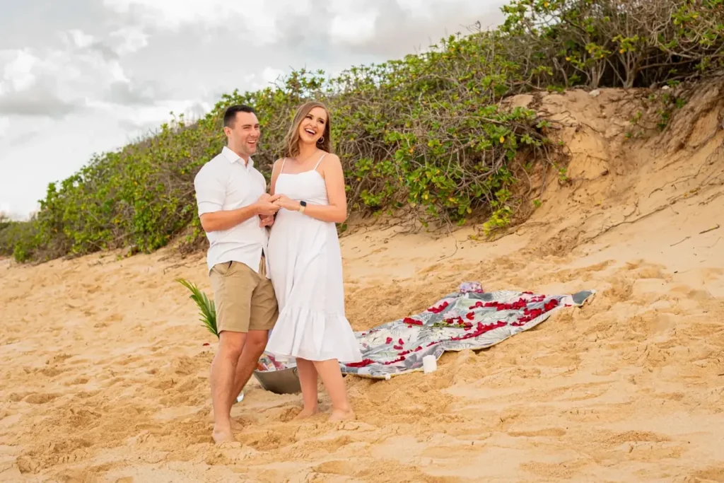 Just-engaged couple laughing on an Oahu beach with a rose petal proposal setup, photographed by Dreamgraphy