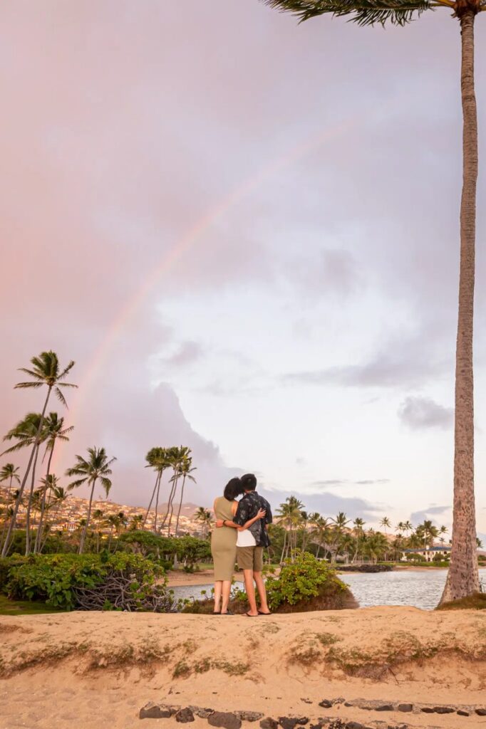 A couple embracing under a rainbow on a beach in Oahu, showcasing a simple and intimate beach proposal setup.