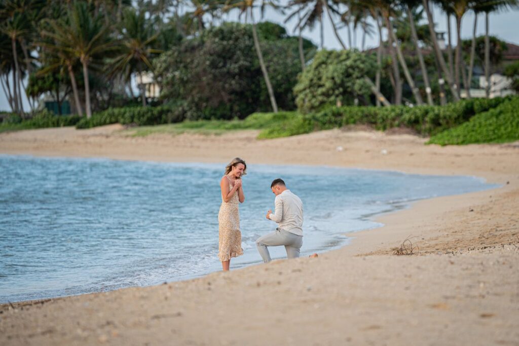 A man proposing on one knee at the waterline during a sunrise beach proposal in Oahu, Hawaii, with palm trees in the background.